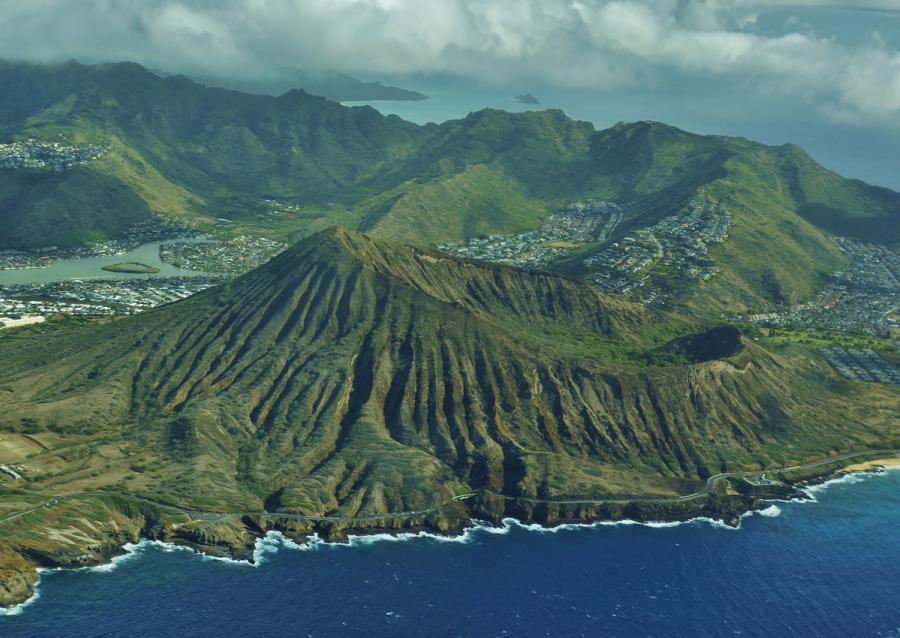 Aerial view of Oahu’s southeast coastline with ridged green mountains, a coastal road, nearby neighborhoods, and waves breaking along the shore.
