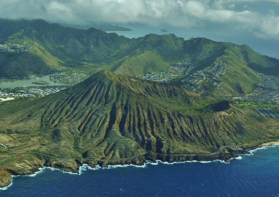 Aerial view of Oahu’s southeast coastline with ridged green mountains, a coastal road, nearby neighborhoods, and waves breaking along the shore.