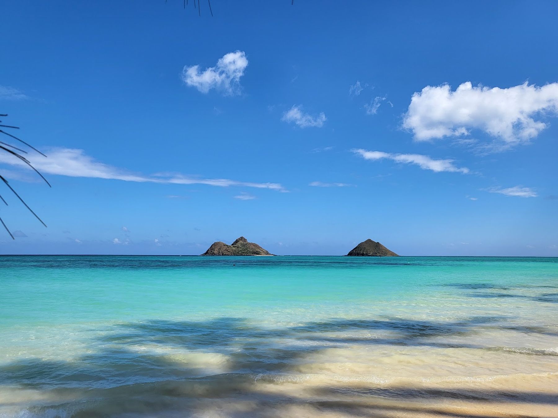 The Mokulua Islands offshore from Lanikai Beach, with clear turquoise water and pale sand in the foreground.