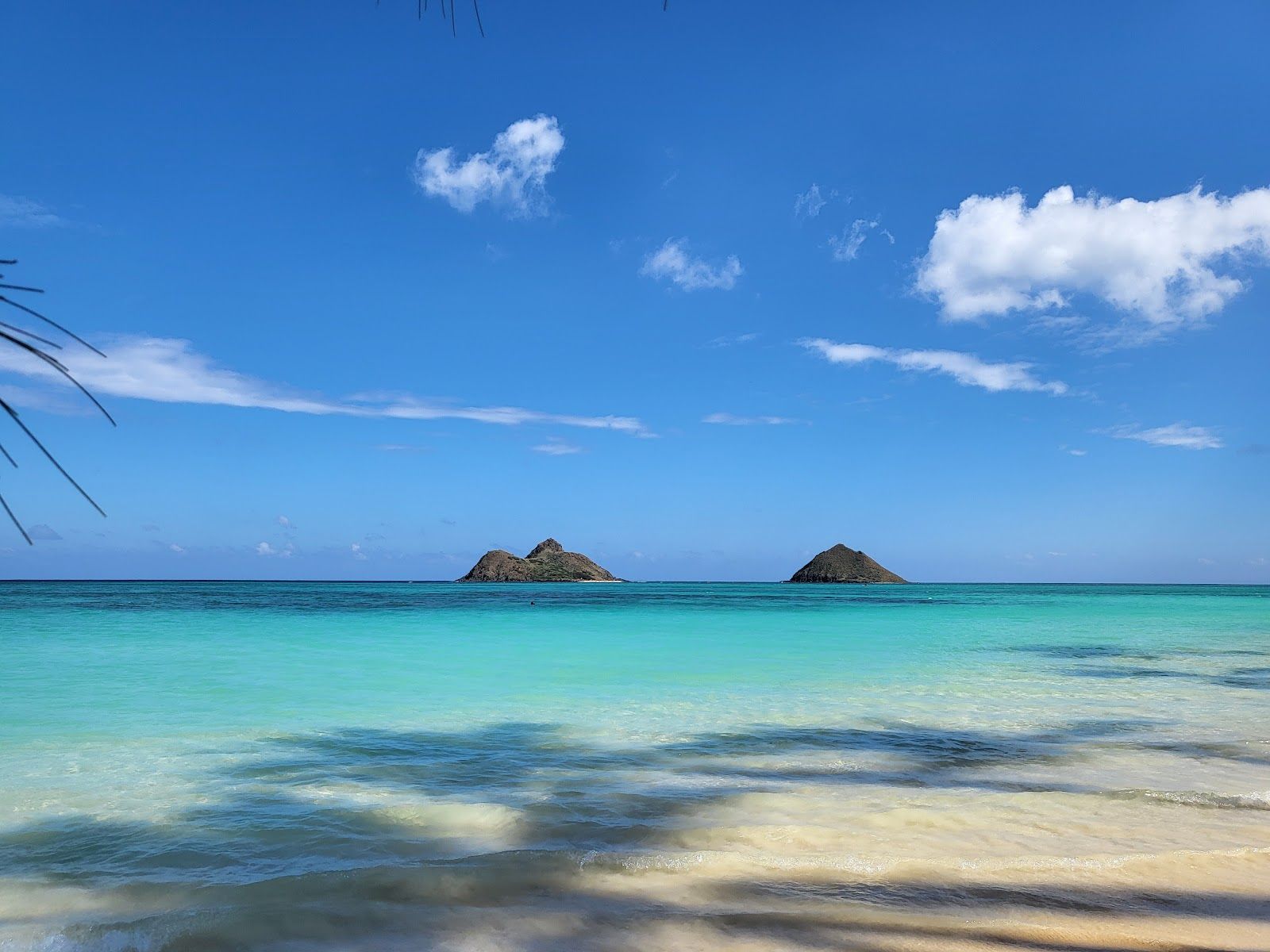 The Mokulua Islands offshore from Lanikai Beach, with clear turquoise water and pale sand in the foreground.