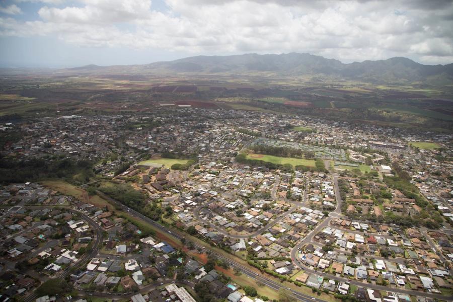 Aerial view of Mililani, Oahu, showing suburban neighborhoods, parks, and roads with farm fields and mountains under a cloudy sky.