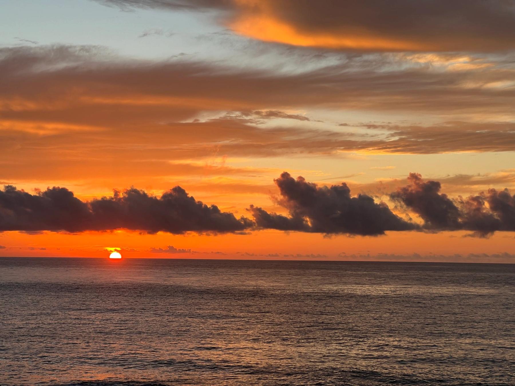 Sun setting over the Pacific with orange and gold clouds above a calm, darkening ocean horizon