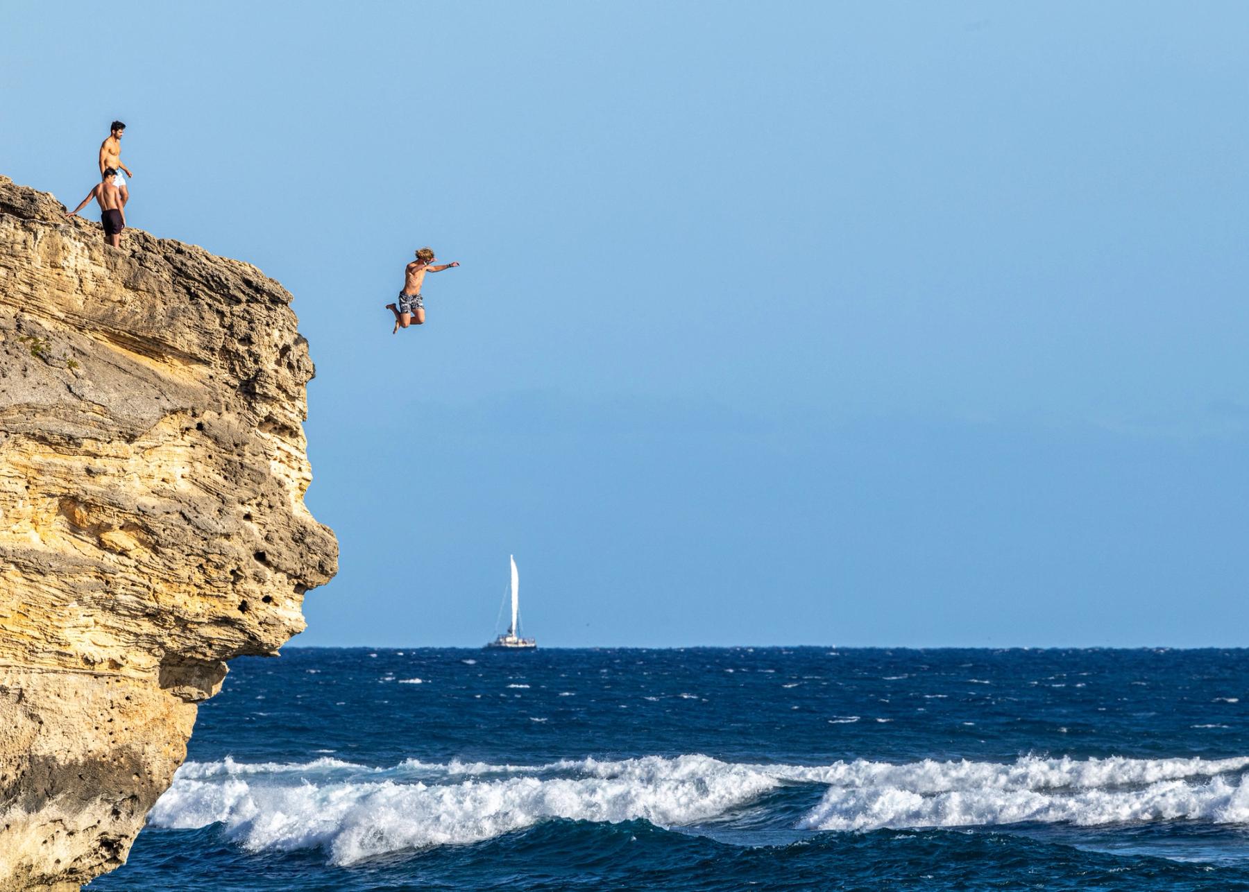 Person midair jumping off a rugged seaside cliff above choppy blue waves, with a sailboat on the horizon under a clear sky.