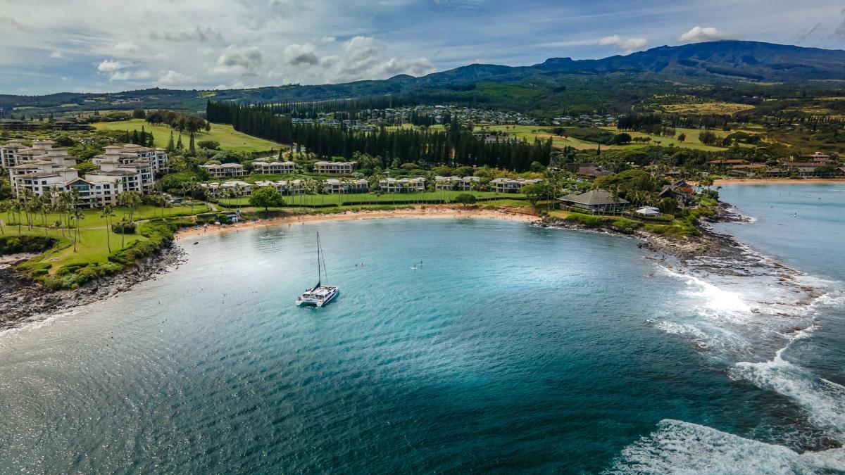 Aerial view of a sailboat in a sheltered bay with a sandy beach, resort buildings, and green hills in West Maui.