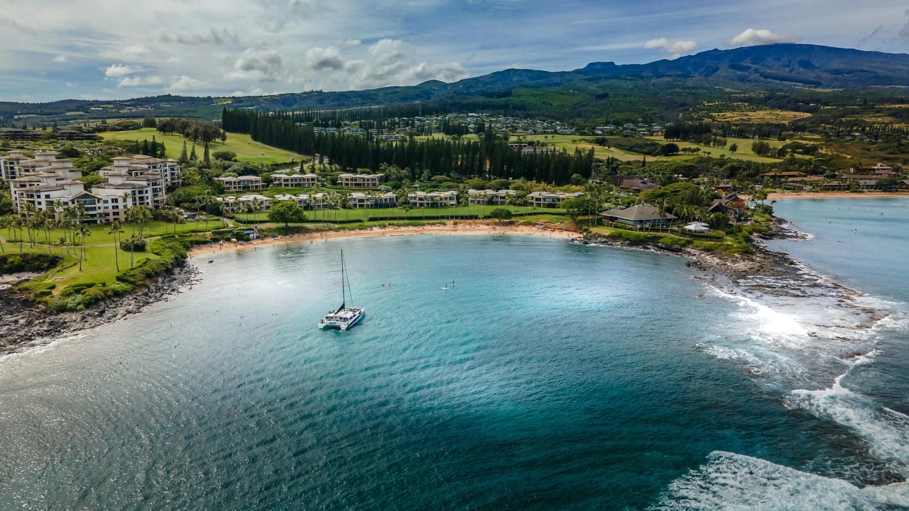 Aerial view of a sailboat in a sheltered bay with a sandy beach, resort buildings, and green hills in West Maui.