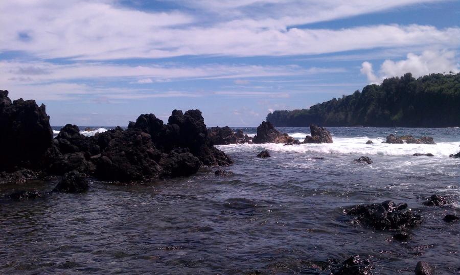 Black lava rocks along the Laupāhoehoe shoreline with waves breaking in the ocean and a forested headland under a partly cloudy sky.