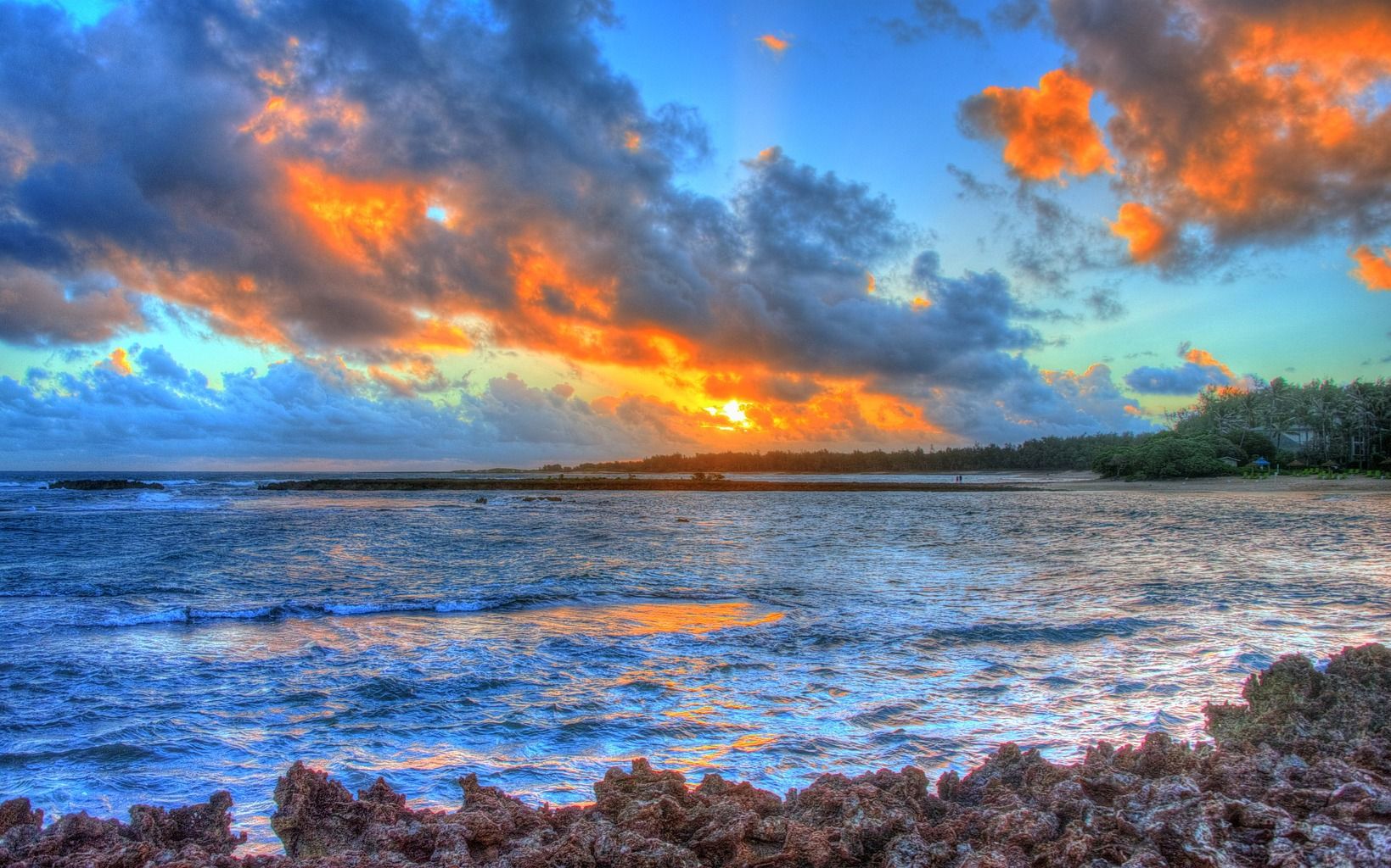 Sunset over the ocean with waves breaking near a rocky shoreline and a palm-lined beach in the distance on Oahu’s North Shore.