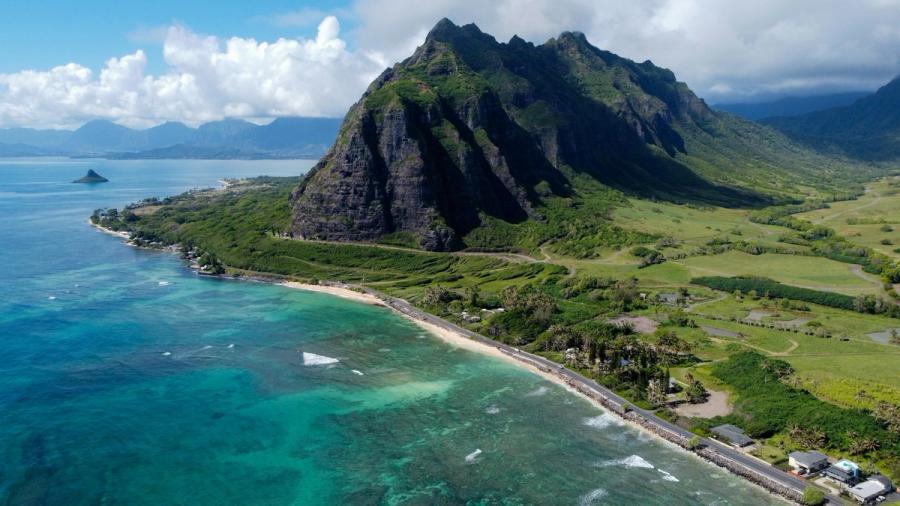 Aerial view of turquoise ocean along a narrow beach and coastal road beneath steep green mountains on Oʻahu’s Windward Coast.