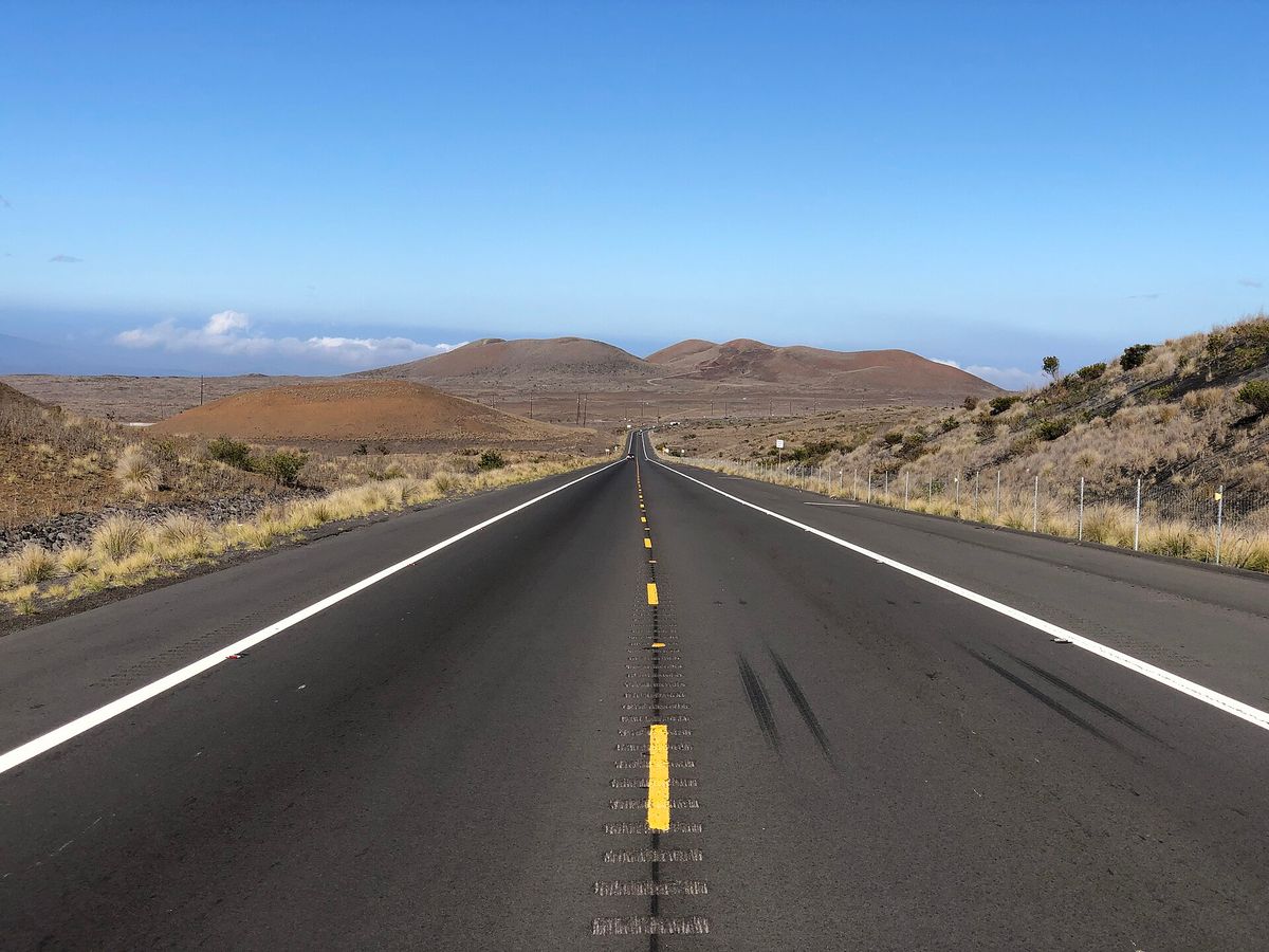 Two-lane Saddle Road stretches through a dry, grassy lava landscape toward low volcanic hills on Hawaii’s Big Island under a clear blue sky.
