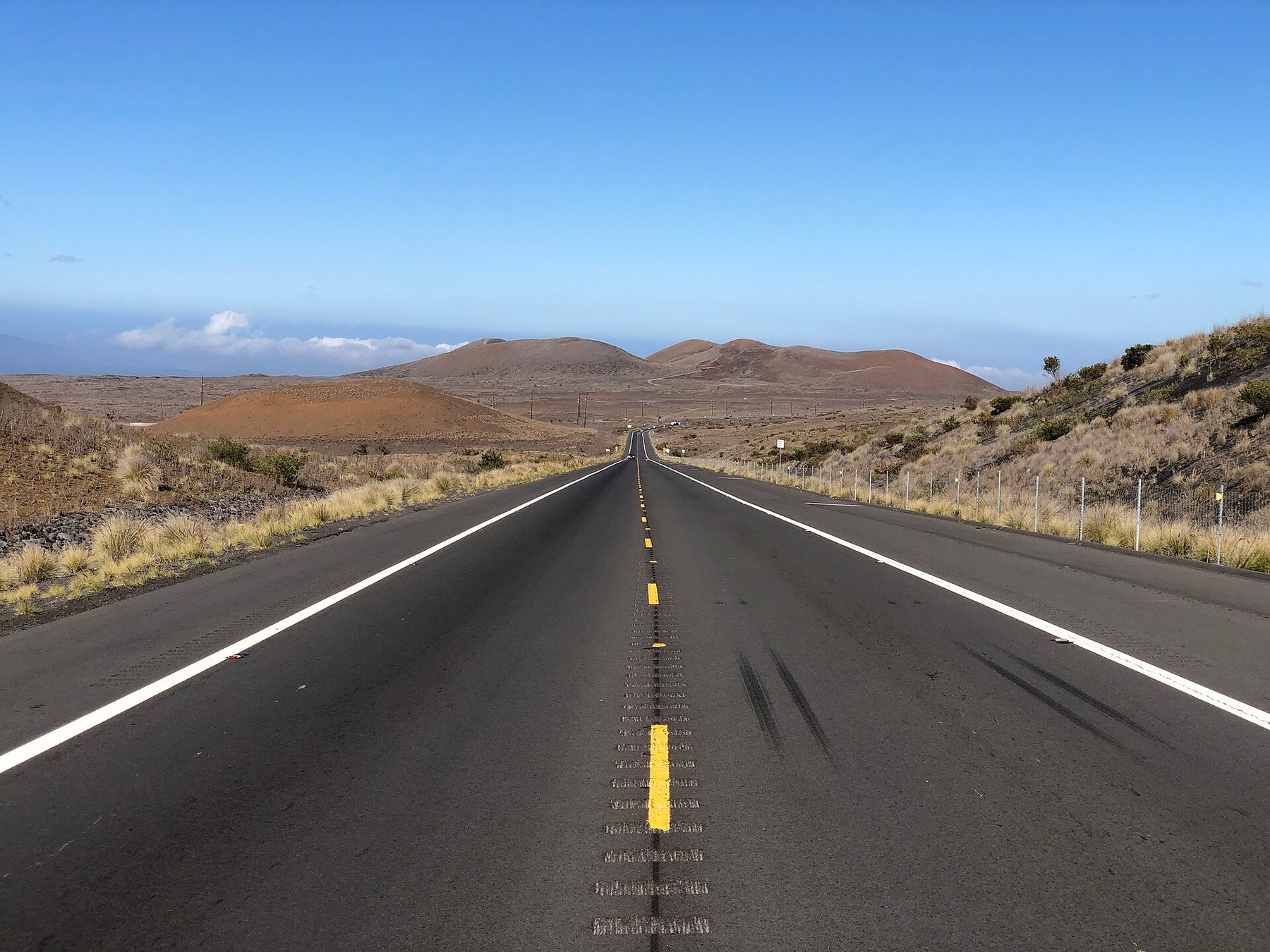 Two-lane Saddle Road stretches through a dry, grassy lava landscape toward low volcanic hills on Hawaii’s Big Island under a clear blue sky.