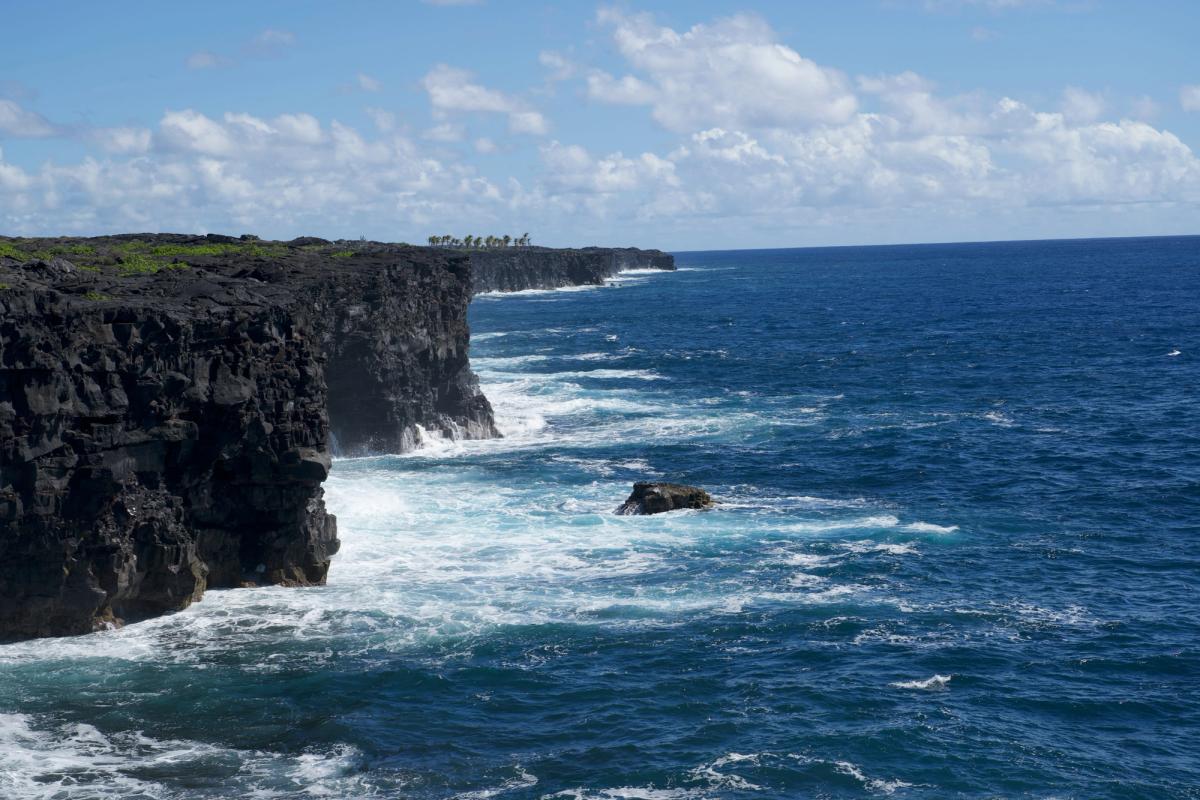 Waves crash against black lava cliffs along the Kaʻū coastline on Hawaiʻi Island under a blue sky with scattered clouds.