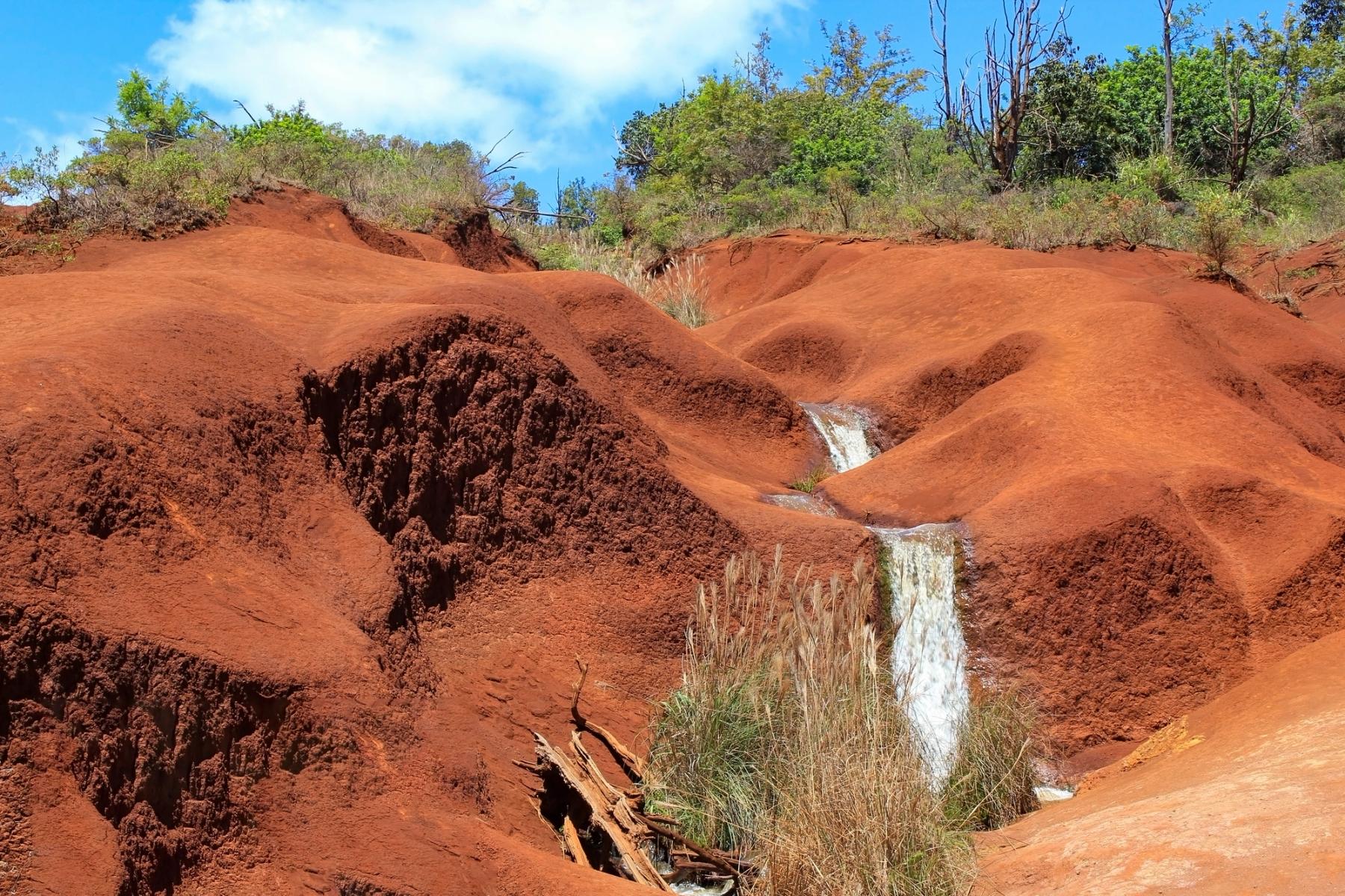 Small stream cascades through smooth red clay hills with green shrubs under a partly cloudy blue sky