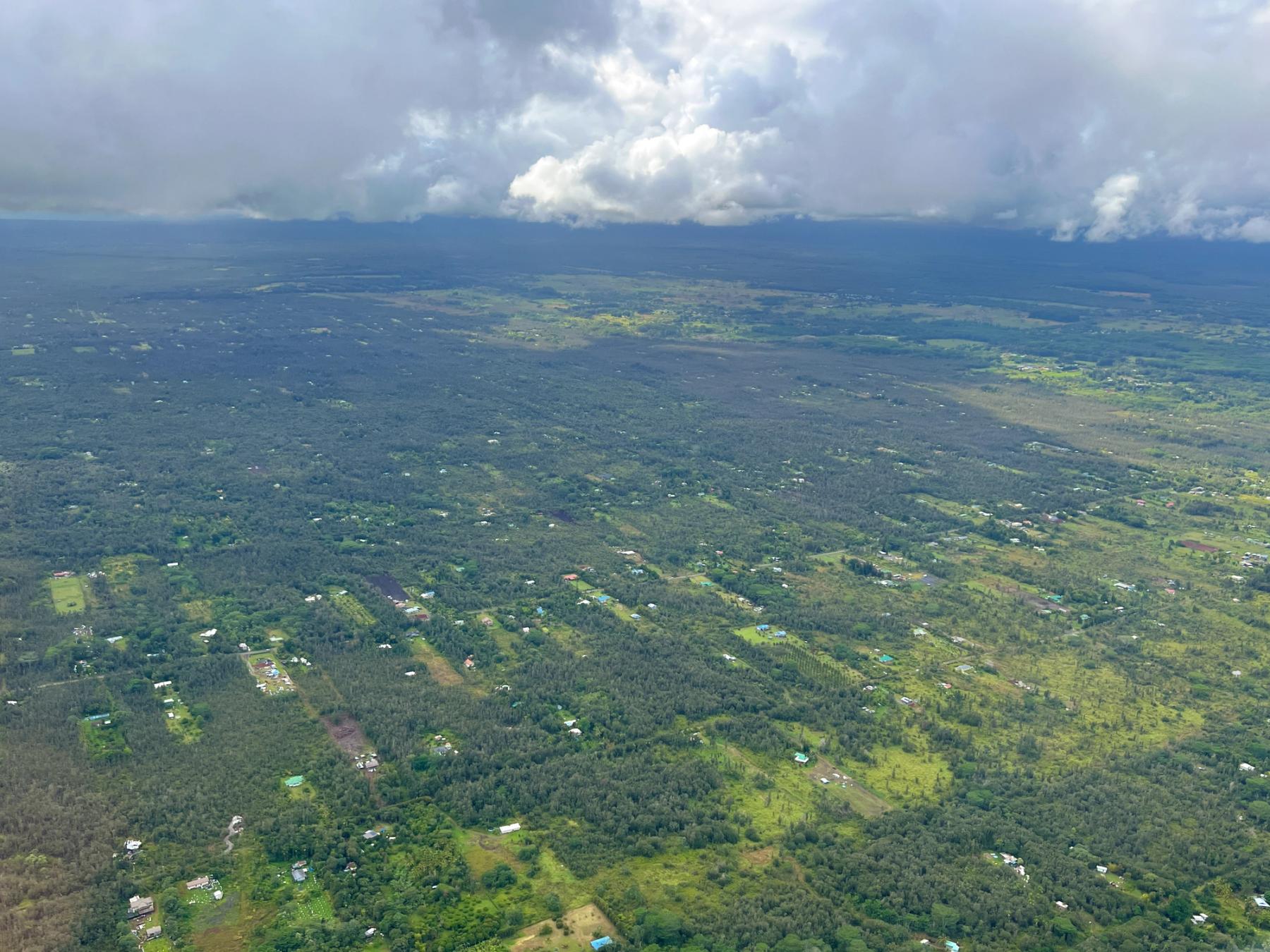 Aerial view of scattered homes and green forest across a broad plain beneath dark clouds and distant hills on Hawaii Island.