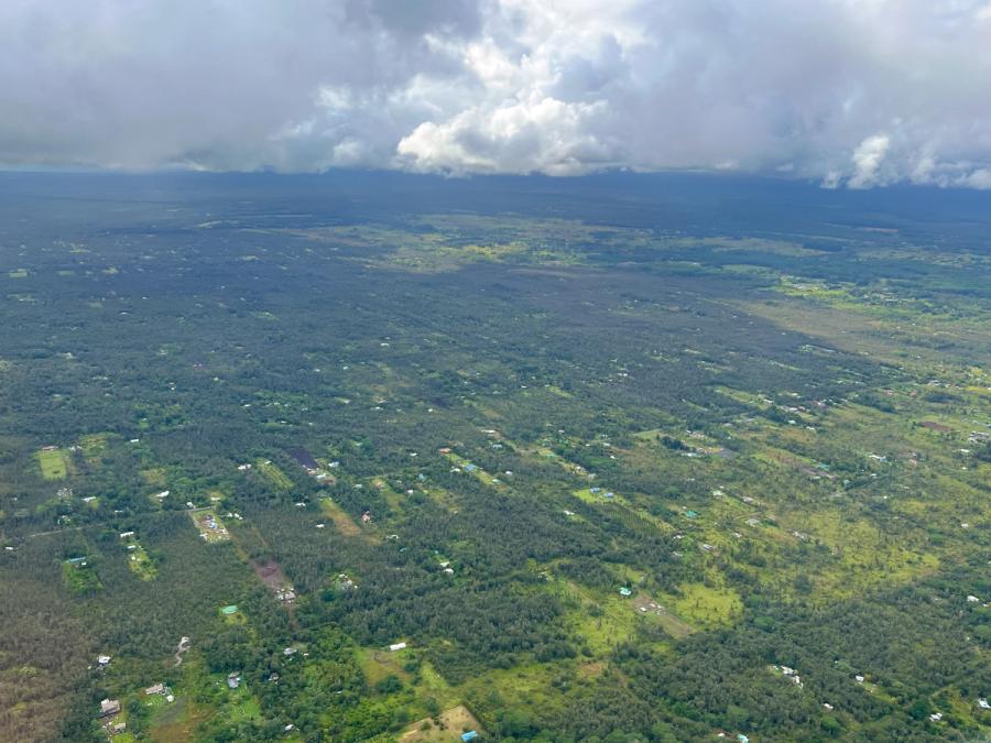 Aerial view of scattered homes and green forest across a broad plain beneath dark clouds and distant hills on Hawaii Island.