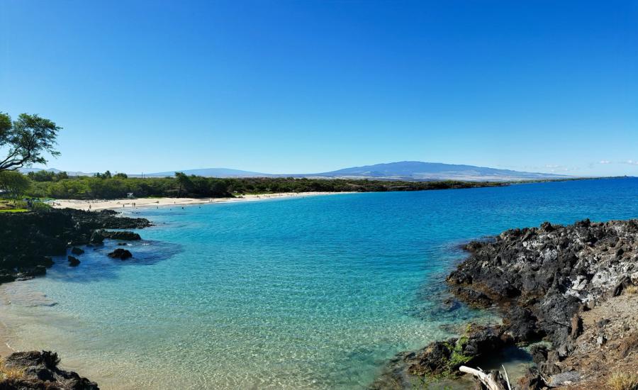 Clear turquoise water along a sandy beach with black lava rocks and distant mountains under a bright blue sky at Mauna Kea Beach on Hawaii Island.