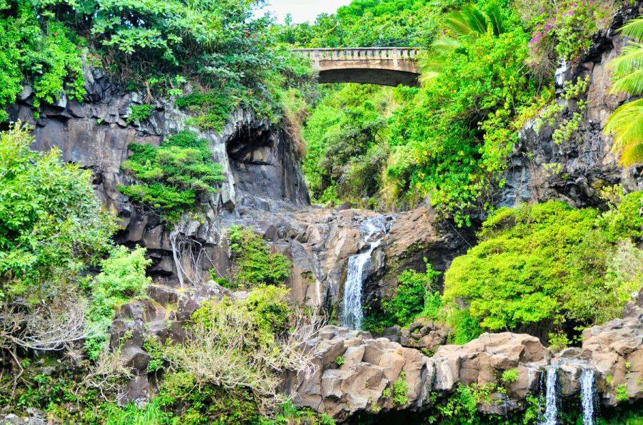 Small waterfalls tumble over rocky cliffs beneath a concrete bridge surrounded by dense green vegetation in Kīpahulu, Maui.