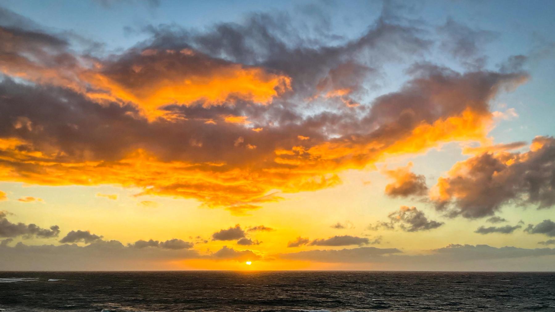 Fiery orange sunrise breaking through clouds above a dark, rippled ocean horizon in Poipū, Kauaʻi
