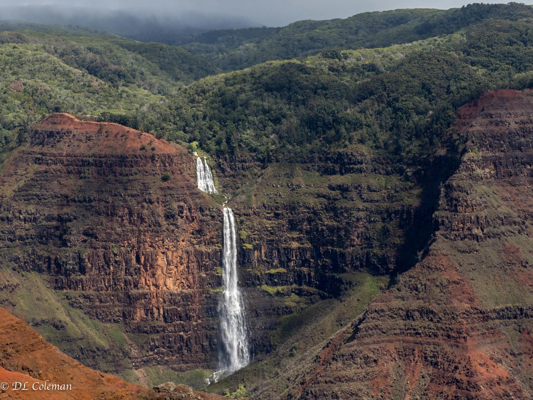 Two-tier Waipoʻo Falls cascading down red and dark basalt cliffs in Waimea Canyon, with green forested ridges and low clouds above.