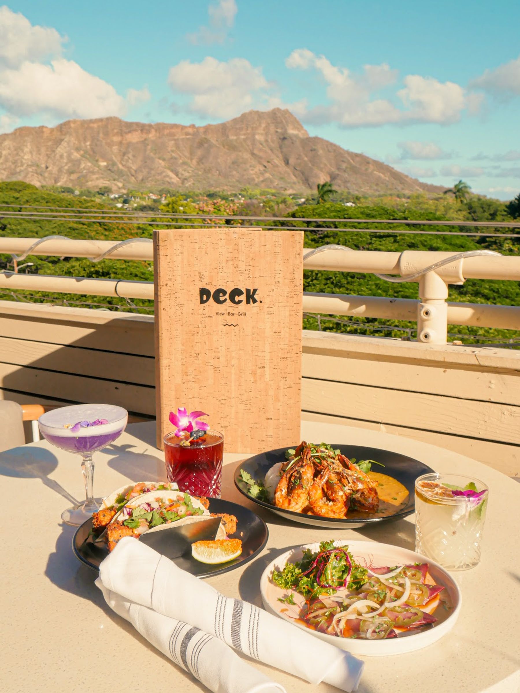 A DECK. menu and colorful dishes set against the Diamond Head view.