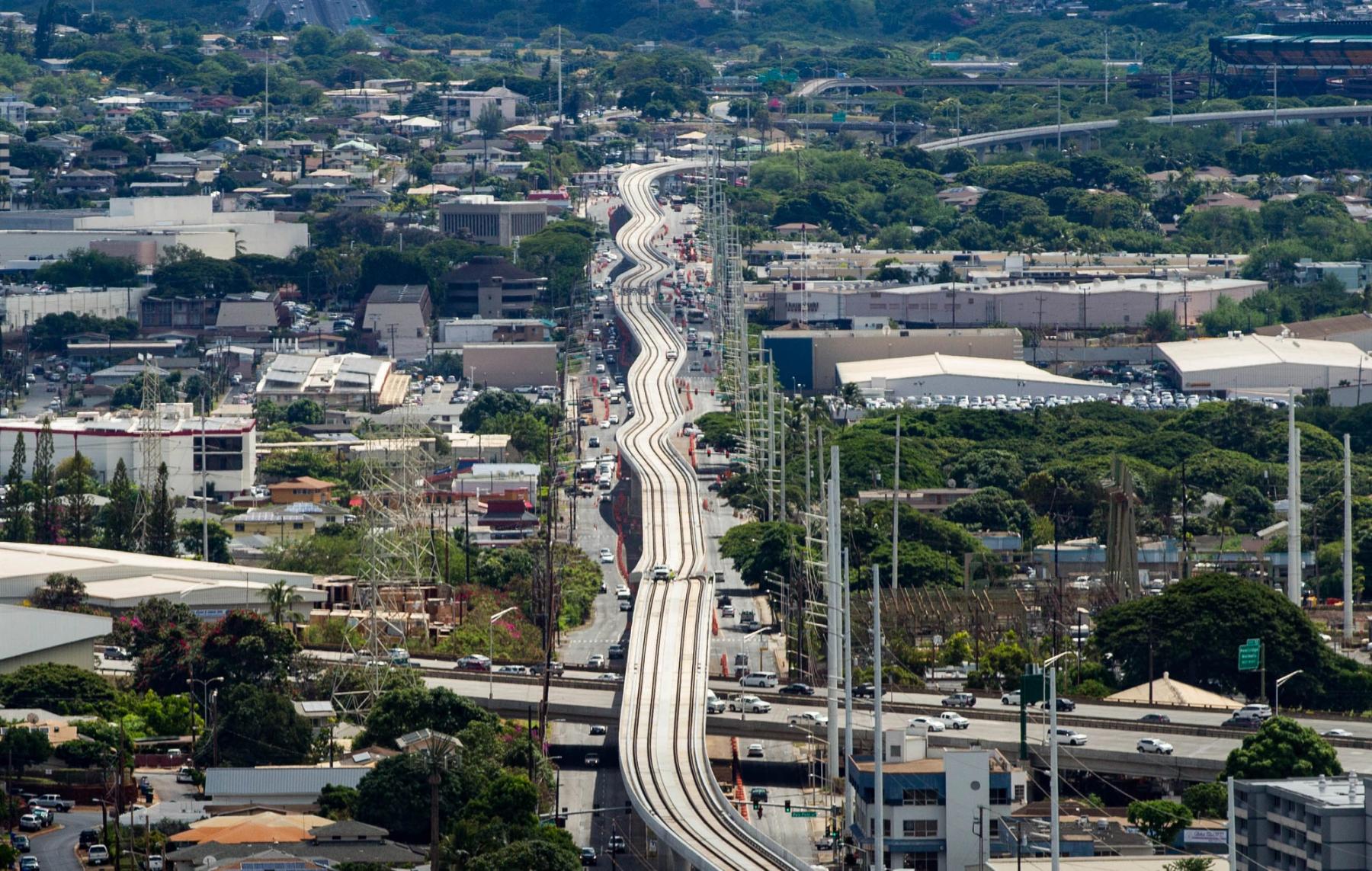 Elevated rail tracks run through Pearl City, Oʻahu, with traffic, warehouses, and tree-covered neighborhoods below.