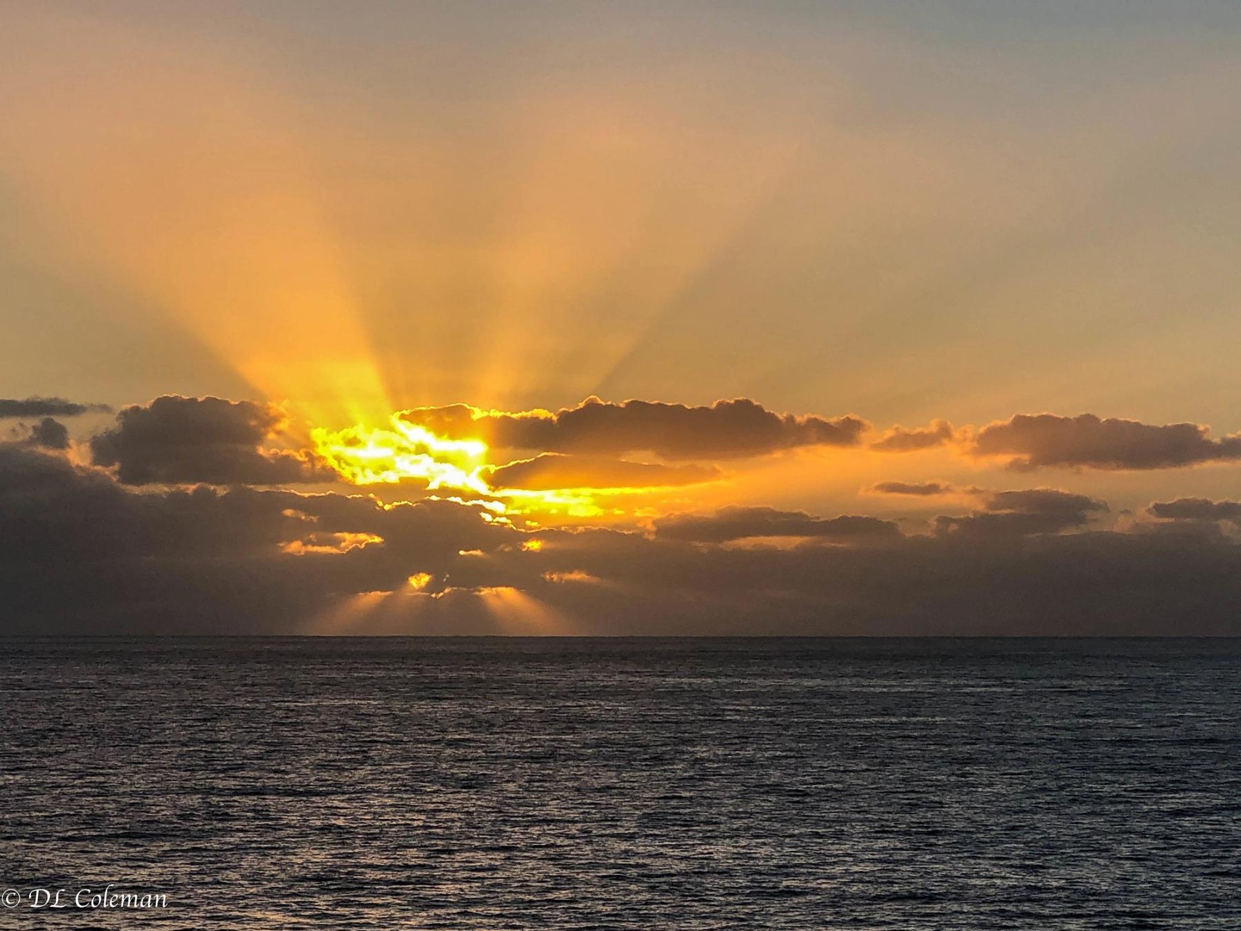 Golden sunrise sunbeams breaking through dark clouds over the ocean horizon near Poʻipū on Kauaʻi