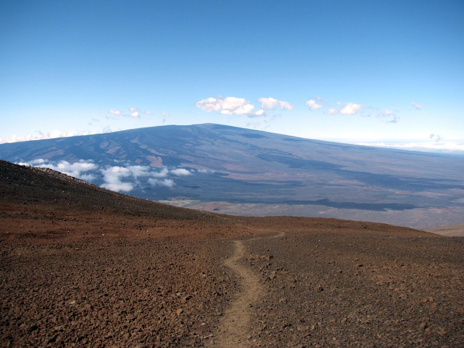 Rocky volcanic trail leading across barren slopes toward Mauna Loa’s broad summit ridge under a clear blue sky with scattered clouds.