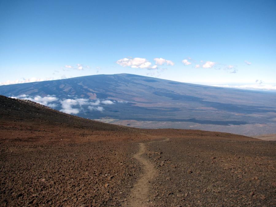 Rocky volcanic trail leading across barren slopes toward Mauna Loa’s broad summit ridge under a clear blue sky with scattered clouds.