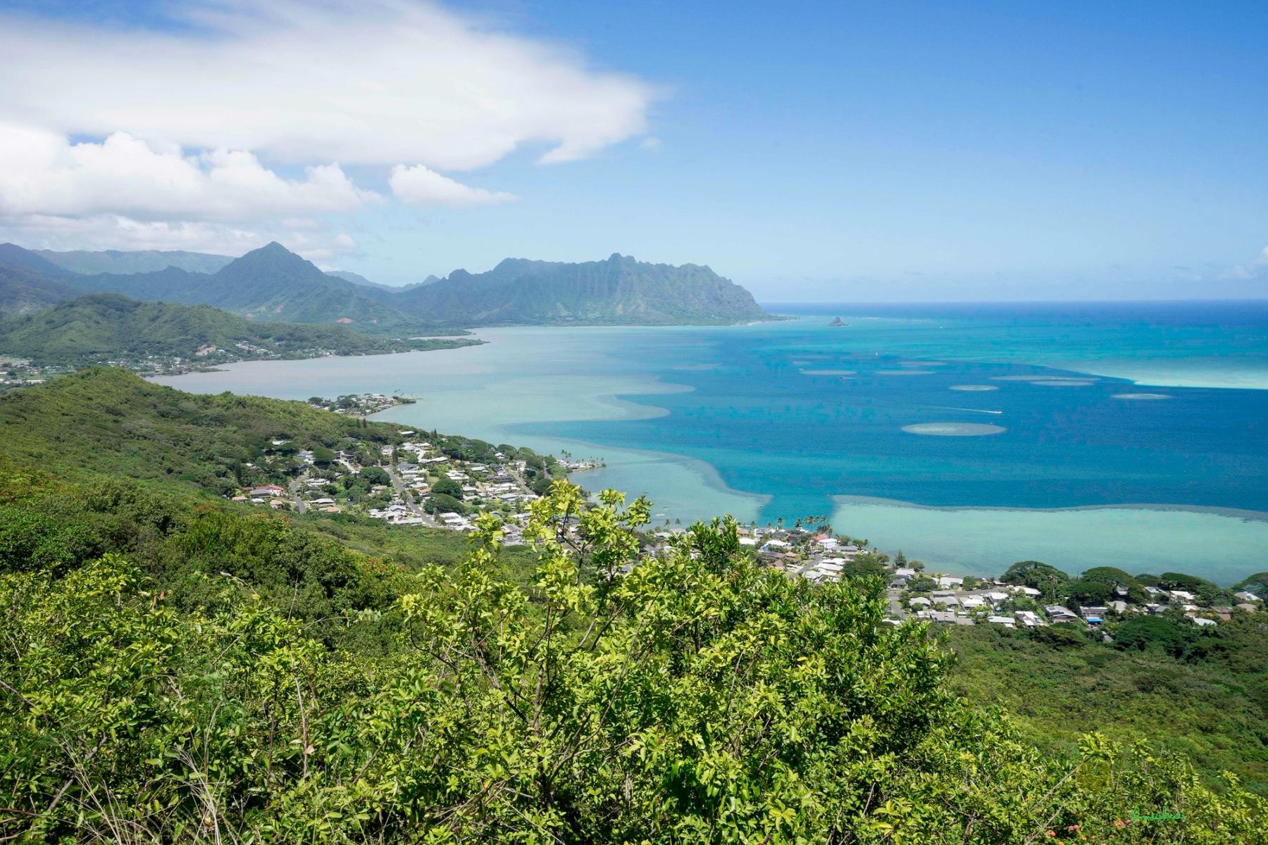 View over Kāneʻohe Bay on Oʻahu with turquoise water, shoreline neighborhoods, and the Koʻolau mountains under a partly cloudy sky.