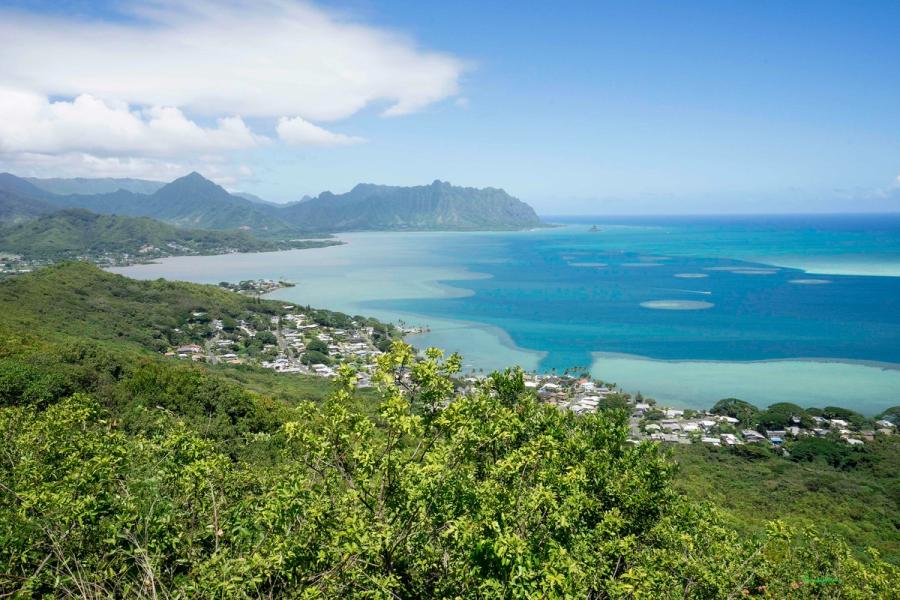 View over Kāneʻohe Bay on Oʻahu with turquoise water, shoreline neighborhoods, and the Koʻolau mountains under a partly cloudy sky.