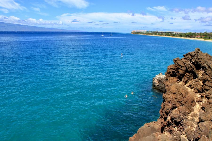 Rocky shoreline overlooks turquoise water with swimmers and sailboats, with Kāʻanapali Beach and resort buildings in the distance under a blue sky.