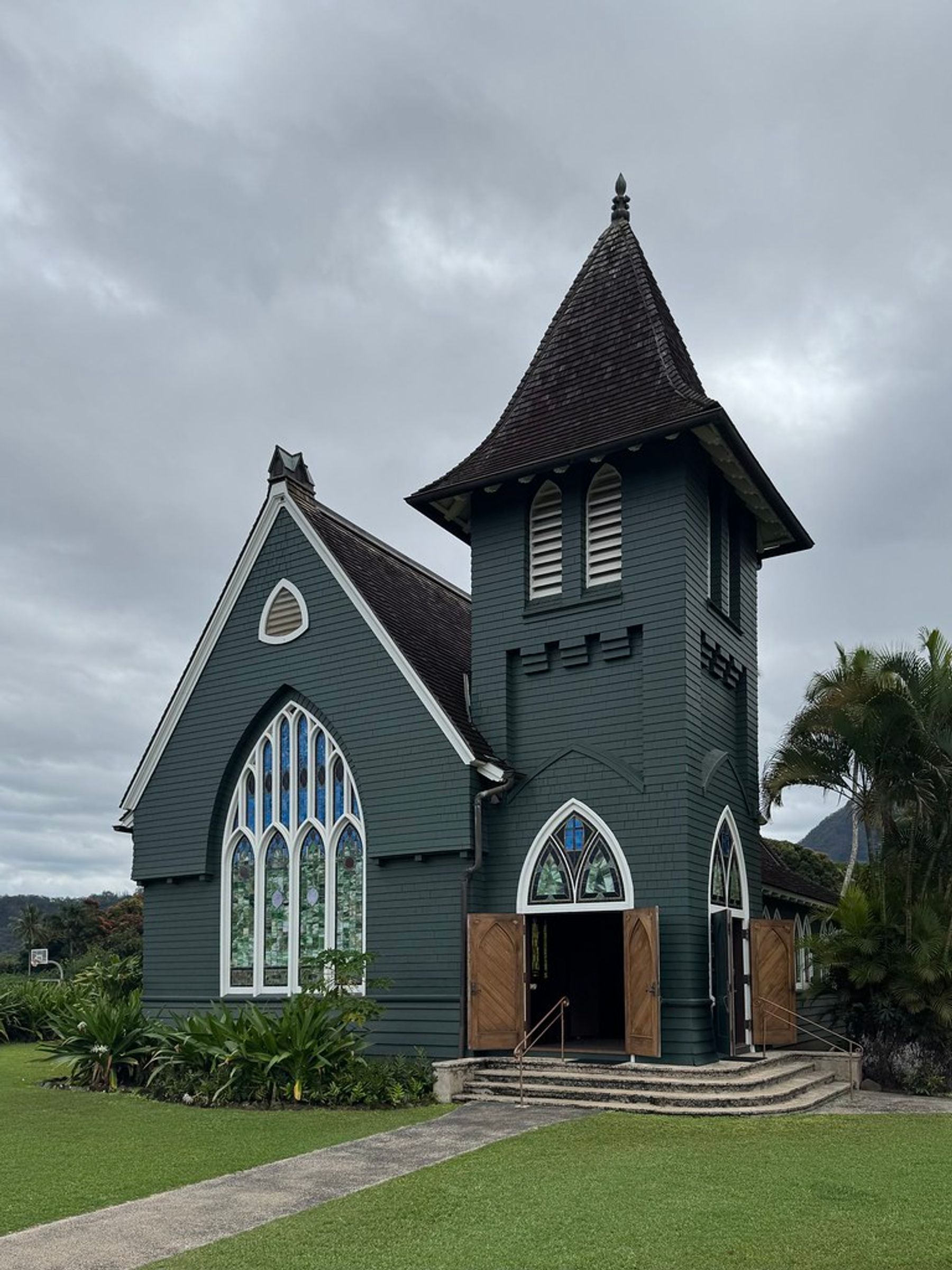 Dark green wooden church with tall tower and arched stained-glass windows under an overcast sky, with a path across a manicured lawn.