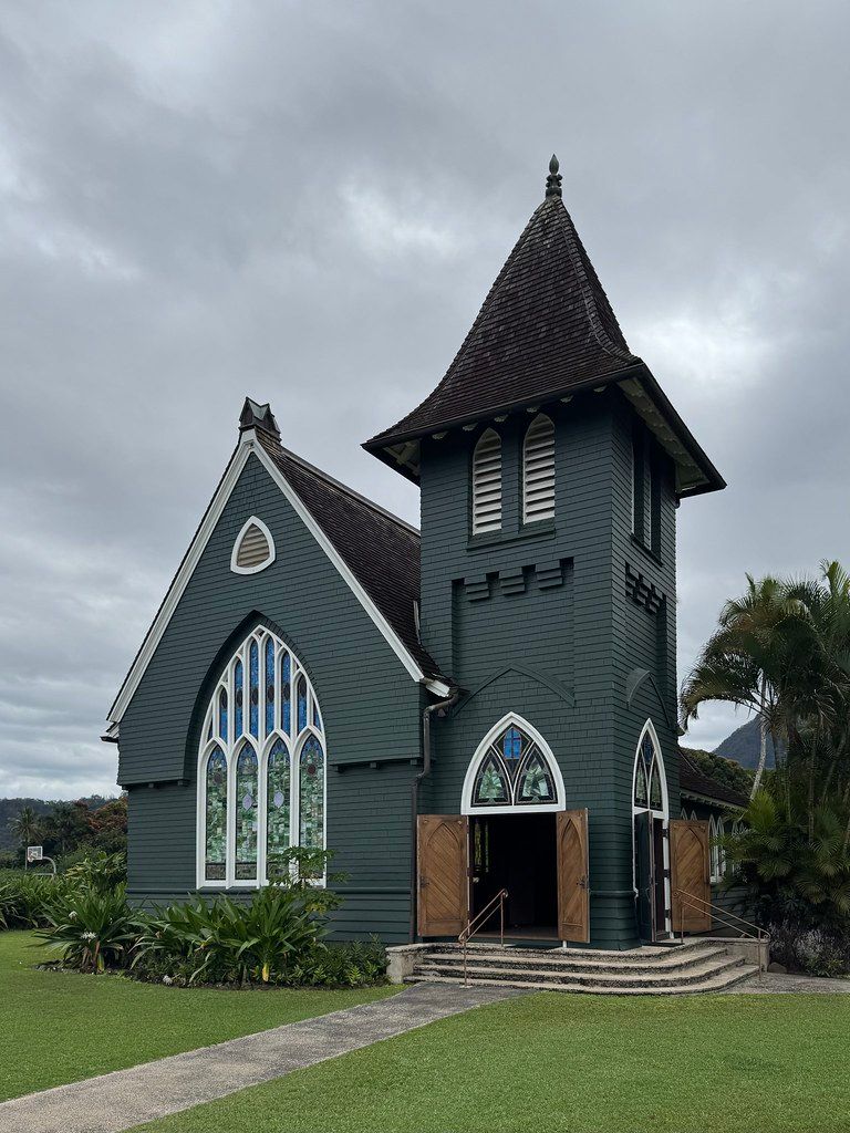 Dark green wooden church with tall tower and arched stained-glass windows under an overcast sky, with a path across a manicured lawn.