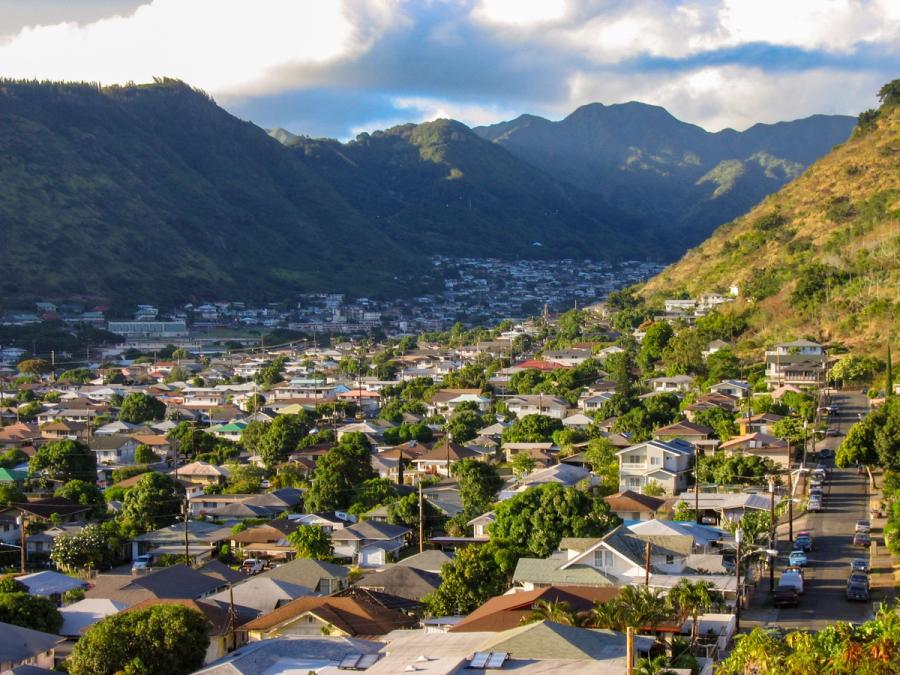 Residential neighborhood rooftops and tree-lined streets in a valley beneath steep green mountains and clouds in Honolulu, Oahu.