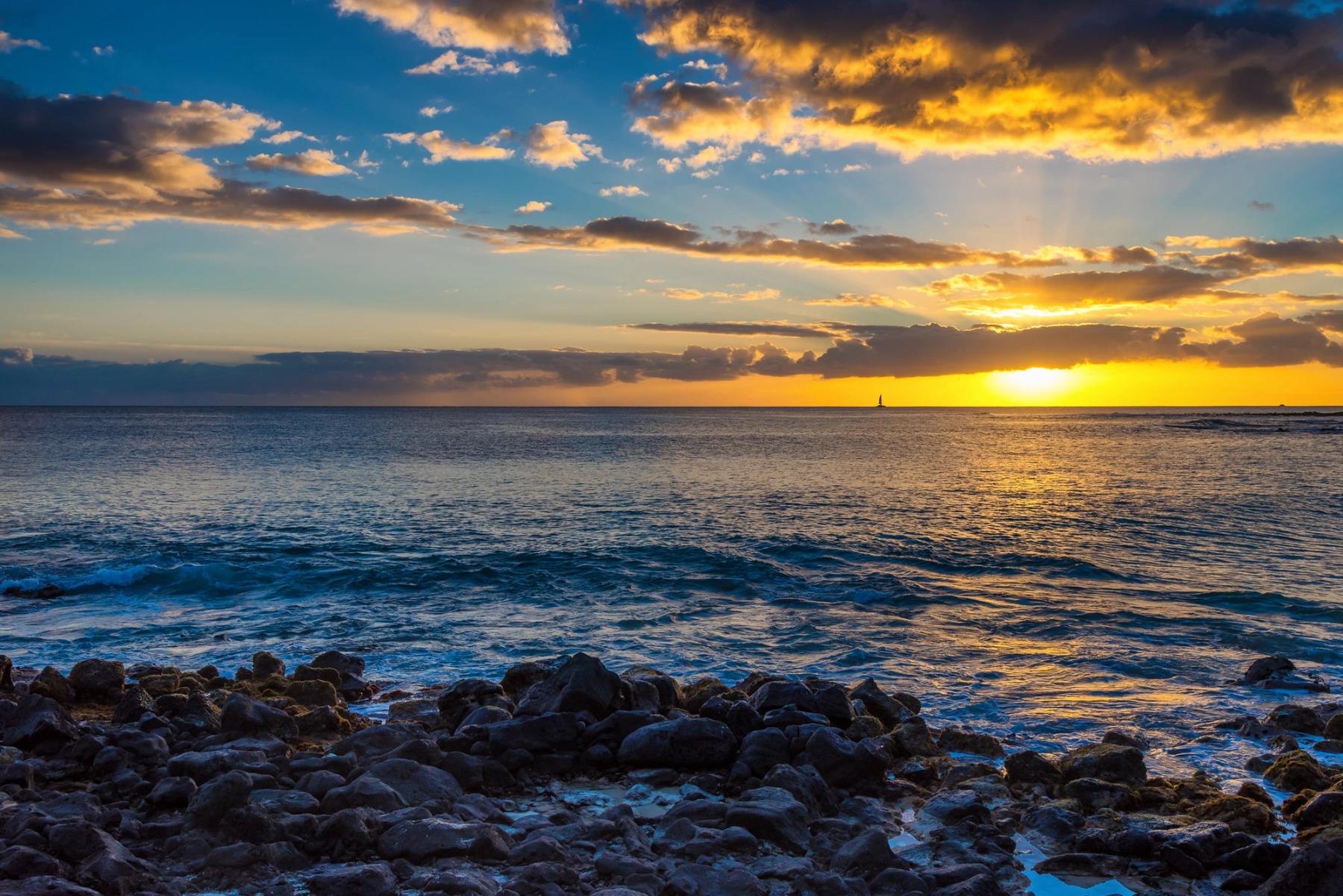 Golden sunset over the ocean with dramatic clouds and dark lava rocks in the foreground on Kauaʻi