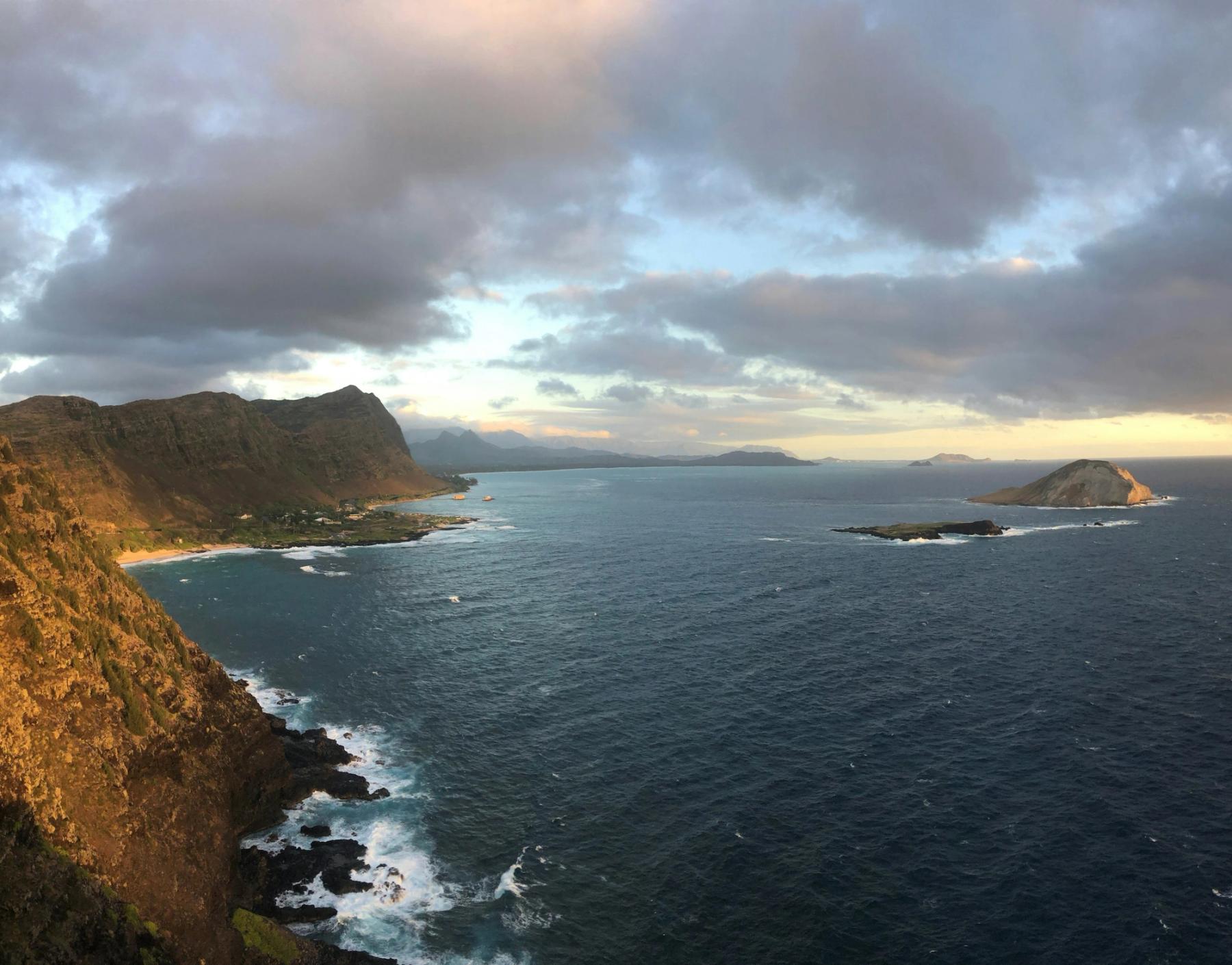 View from a rocky cliff over a curving coastline with distant mountains, choppy ocean, and small offshore islets under a cloudy sky in Waimānalo, Oʻahu.