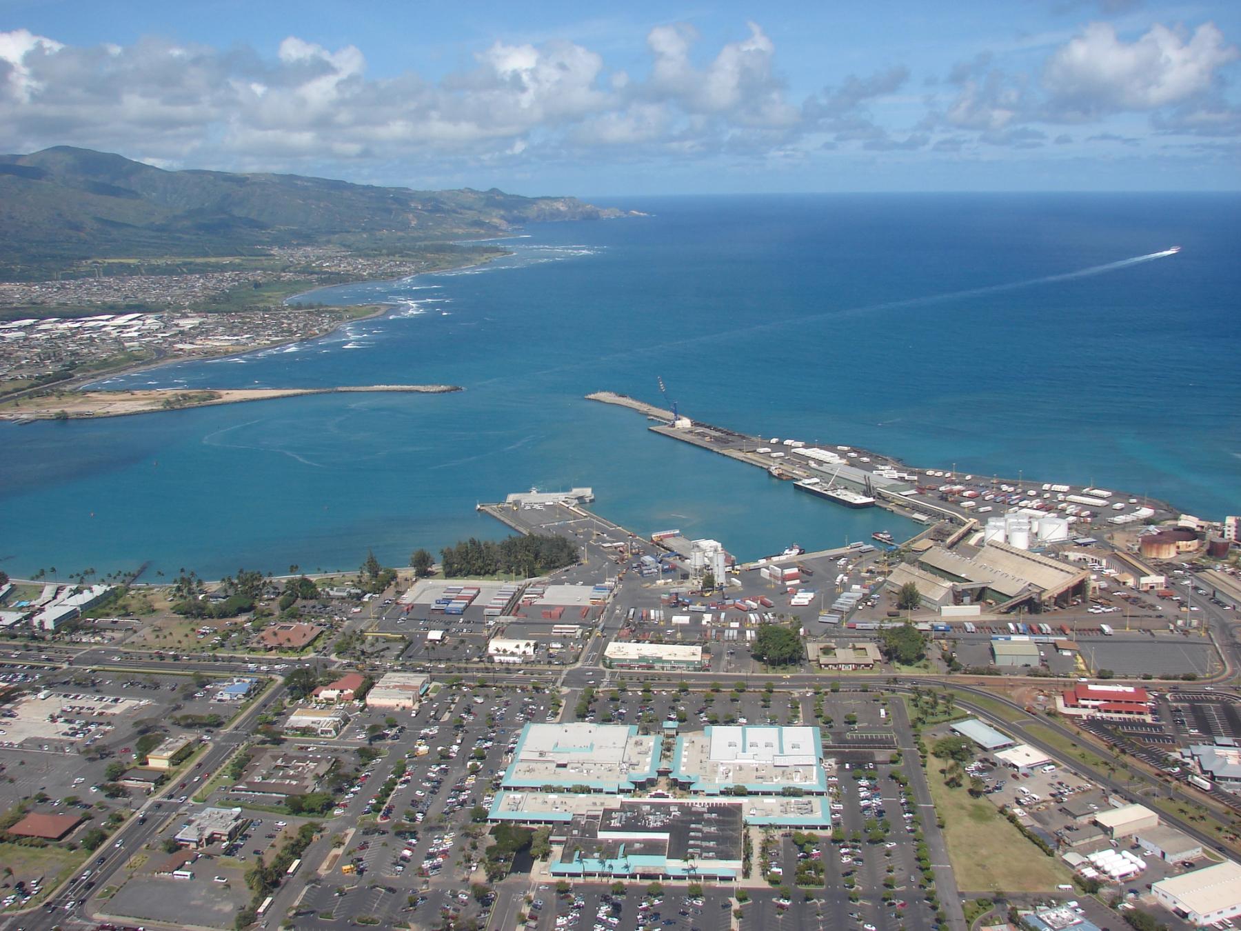 Aerial view of Kahului, Maui, showing the harbor, industrial port facilities, and coastline under a partly cloudy sky.