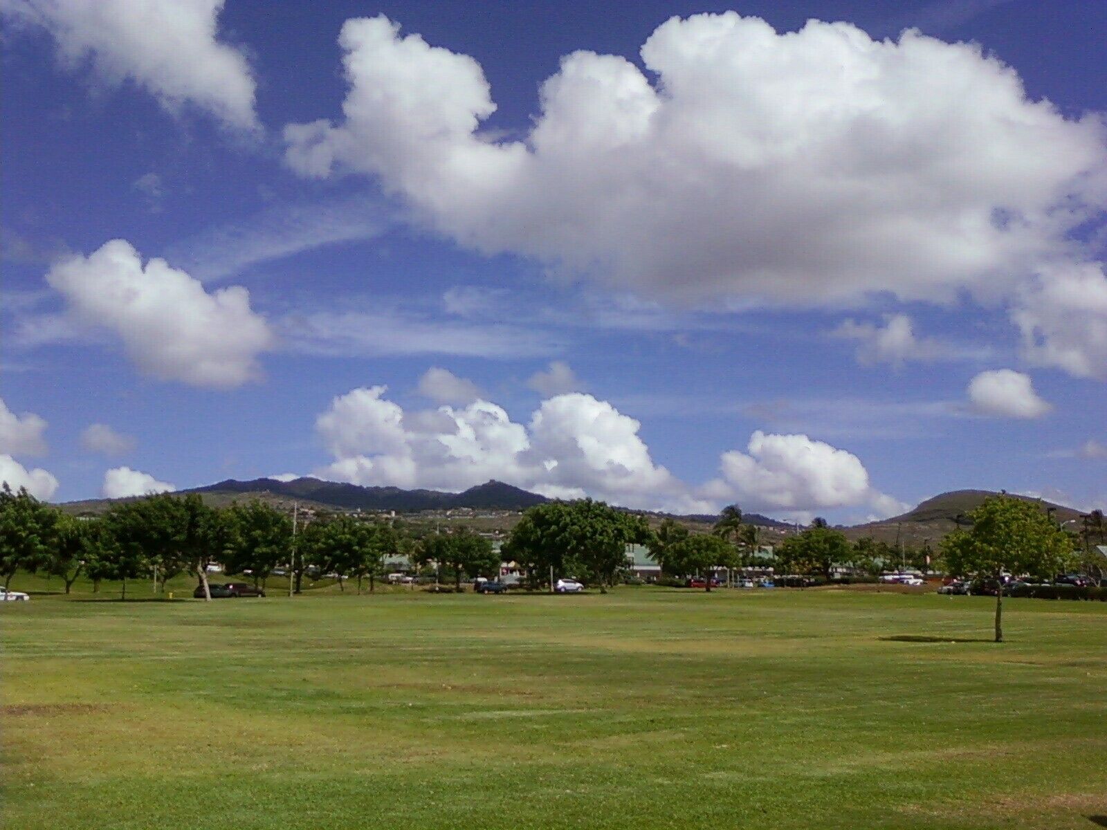 Wide grassy park with scattered trees, parked cars, and low mountains beneath large clouds in Kapolei and Makakilo on Oʻahu.
