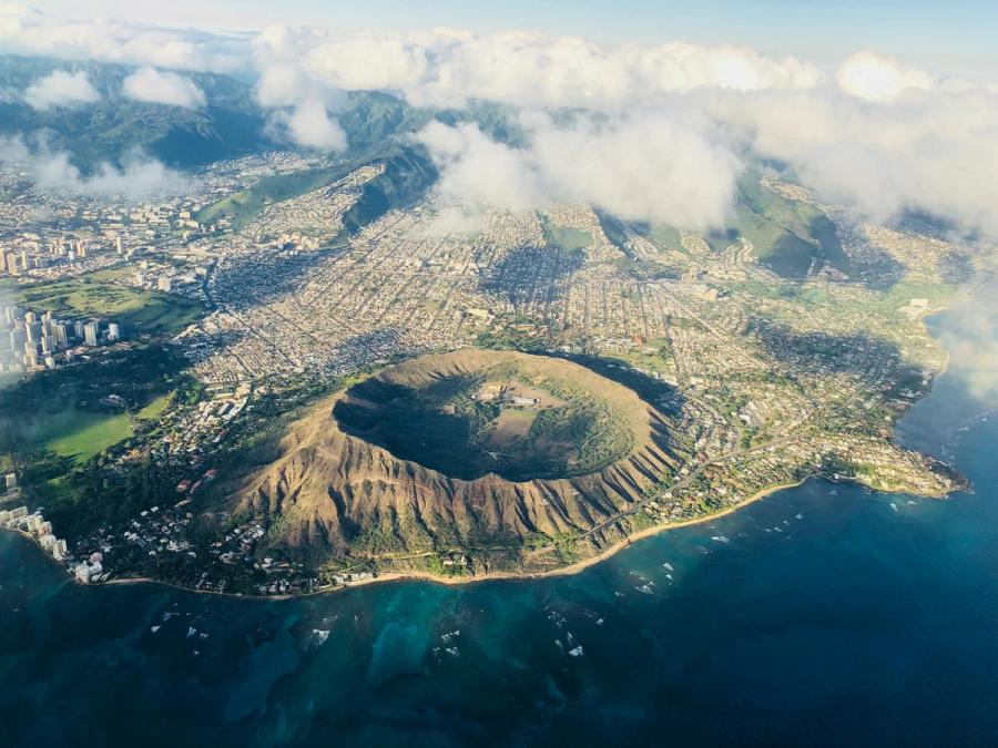 Aerial view of Diamond Head crater on Oahu with Honolulu neighborhoods, shoreline, and scattered clouds over green mountains.