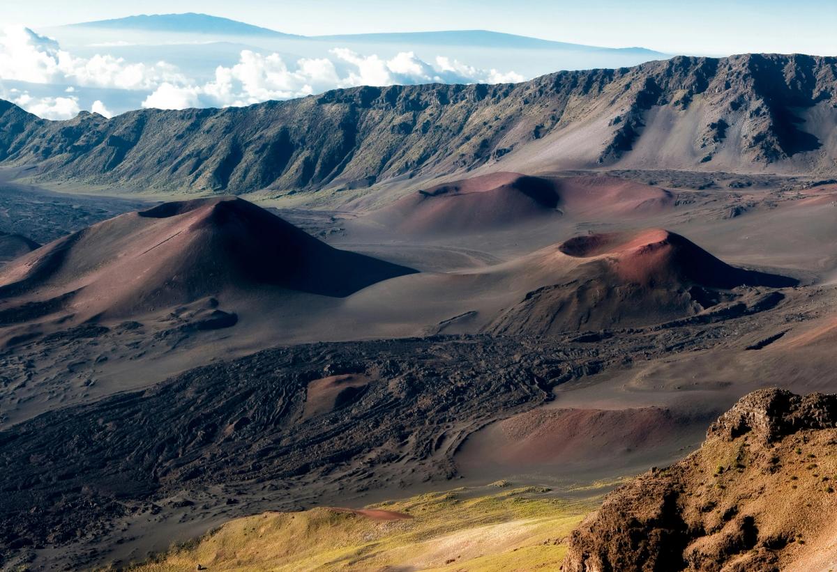 Volcanic cinder cones and dark lava fields inside Haleakalā Crater on Maui, with rocky ridgelines and clouds in the distance.