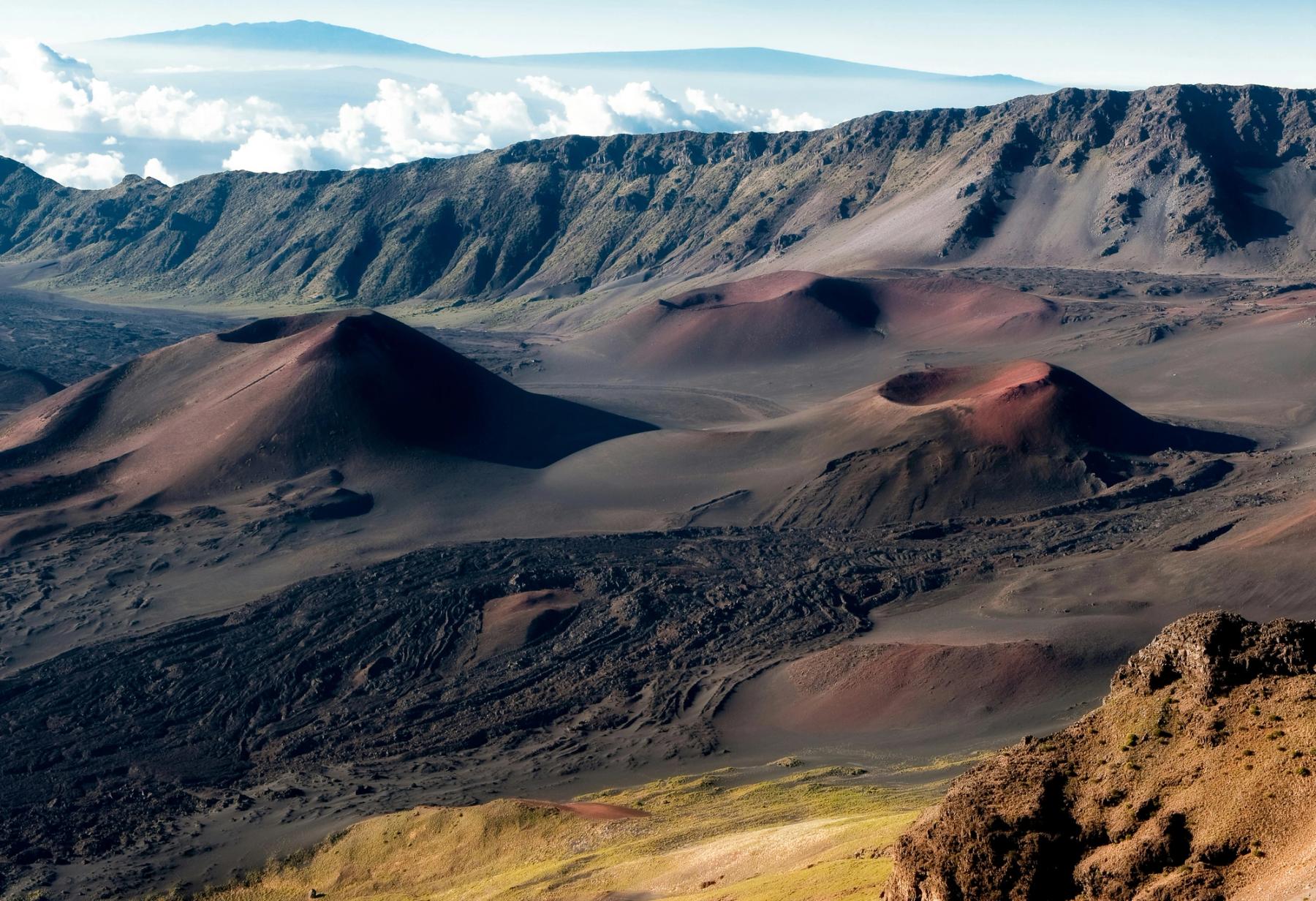 Volcanic cinder cones and dark lava fields inside Haleakalā Crater on Maui, with rocky ridgelines and clouds in the distance.