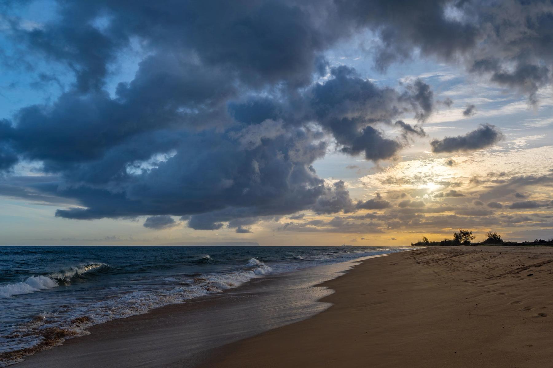 Curving sandy shoreline at Kekaha Beach with small breaking waves and a dramatic cloud-filled sky glowing near sunset
