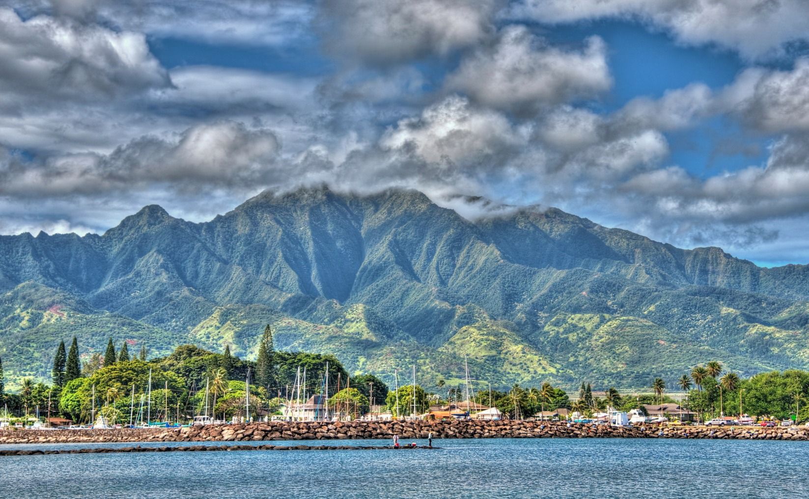 Sailboat masts and a rocky breakwater at Haleʻiwa Harbor sit below green mountains under a cloudy blue sky.