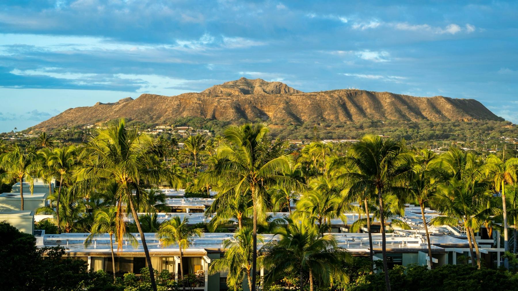 Palm trees and low-rise buildings in the foreground with a sunlit mountain ridge under a partly cloudy sky in Kahala, Oahu.