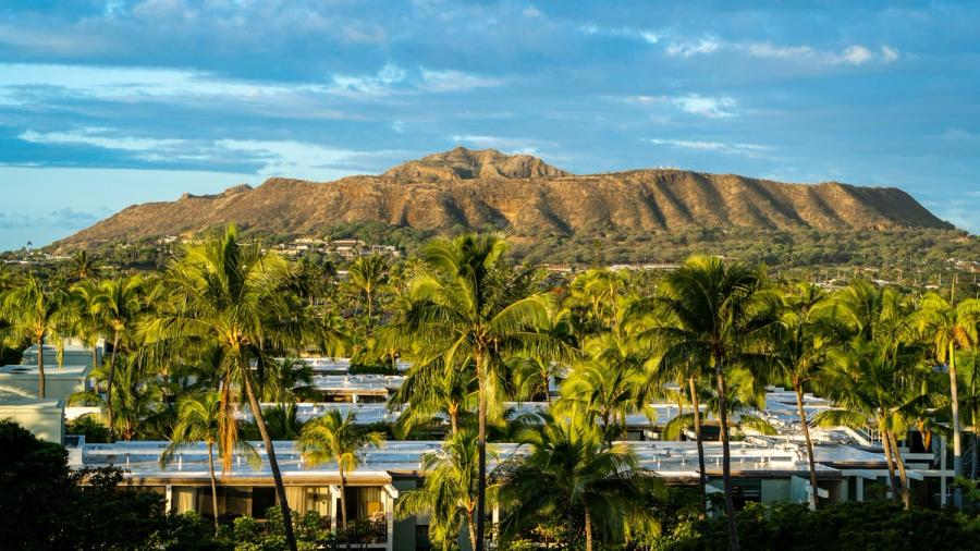Palm trees and low-rise buildings in the foreground with a sunlit mountain ridge under a partly cloudy sky in Kahala, Oahu.