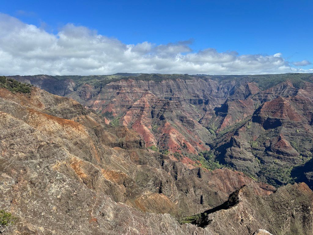 Panoramic view of Waimea Canyon with red and green layered ridges, deep ravines, and scattered clouds under a bright blue sky