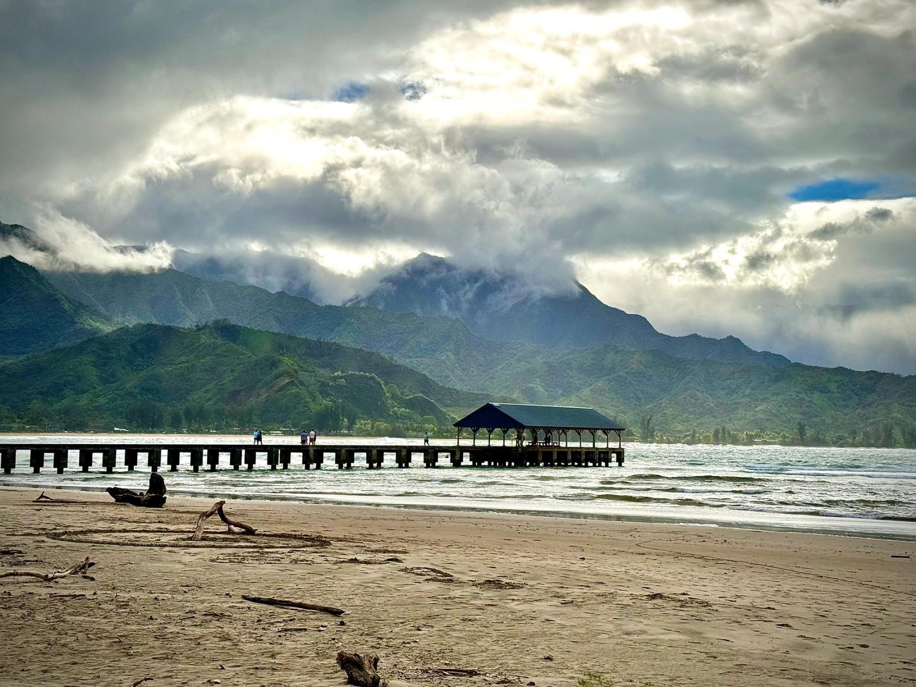 Sandy beach with driftwood in the foreground, a long pier with a pavilion over the water, and lush green mountains under dramatic sunlit clouds.