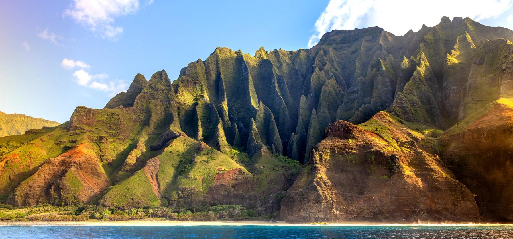 Sunlit Nā Pali Coast cliffs with sharp green ridges above a narrow beach and blue ocean