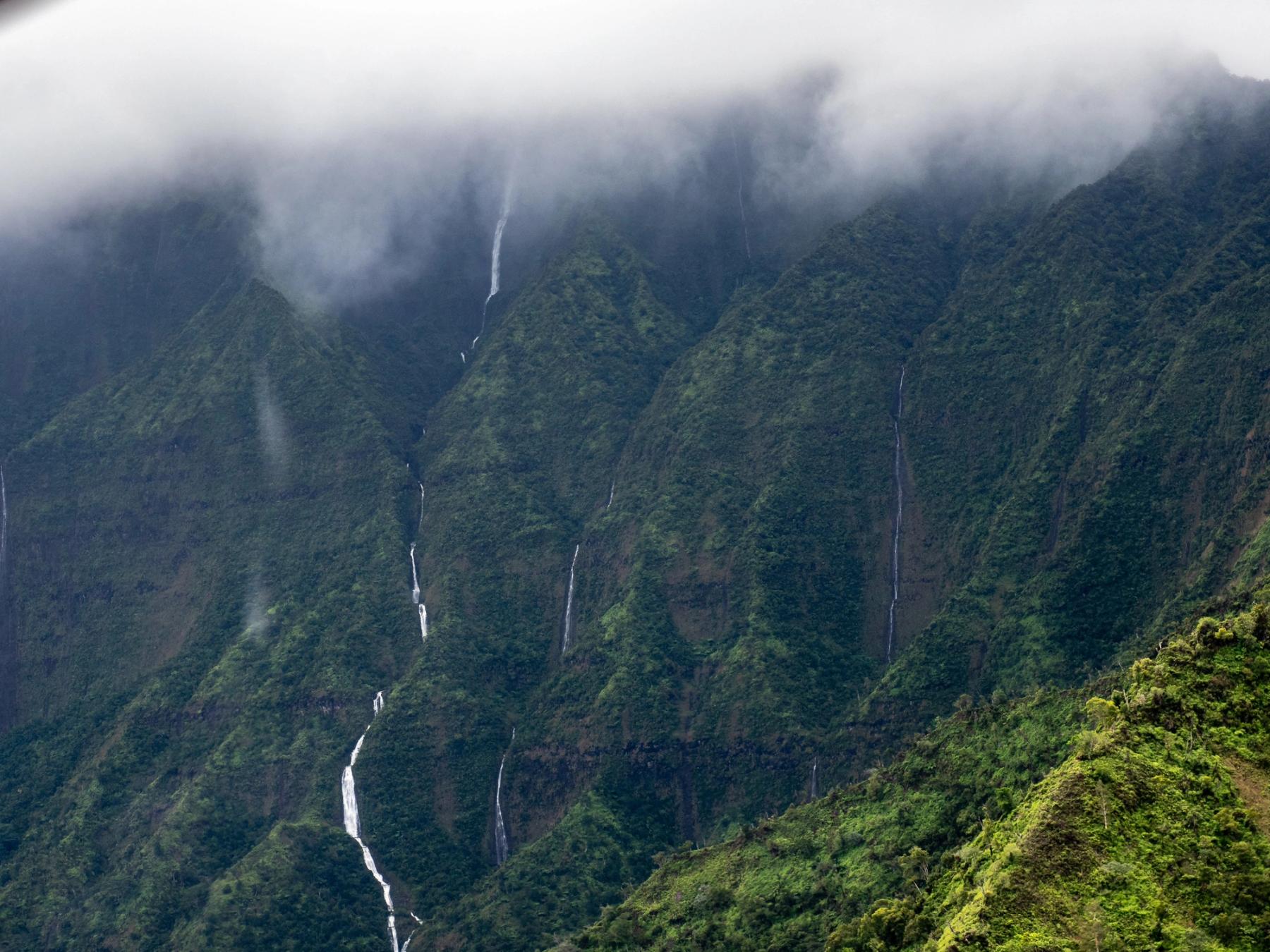 Multiple thin waterfalls spilling down lush green Kauaʻi cliffs beneath low misty clouds