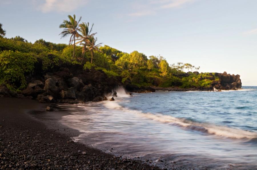 Waves wash onto a black sand beach beneath palm trees and lush greenery along the coastline in Hāna, Maui.