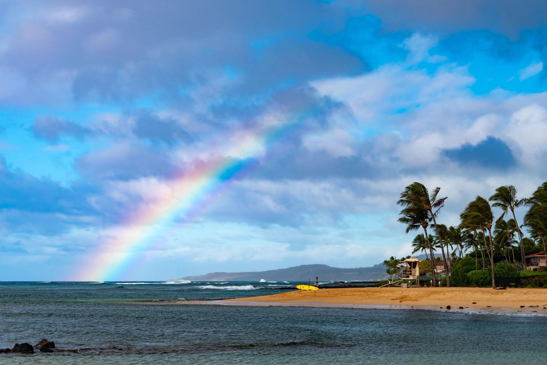 Rainbow over Poʻipū beach with surf, sandy shoreline, and palm trees under dramatic blue-gray clouds